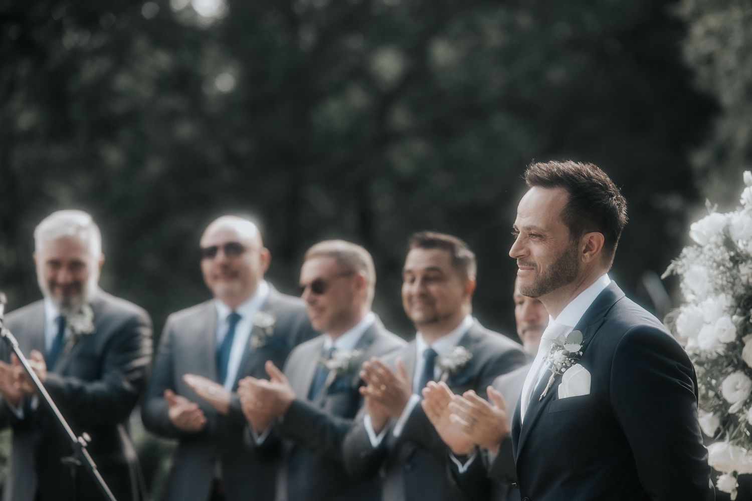 Groomsmen in dark suits stand together during an outdoor wedding ceremony.