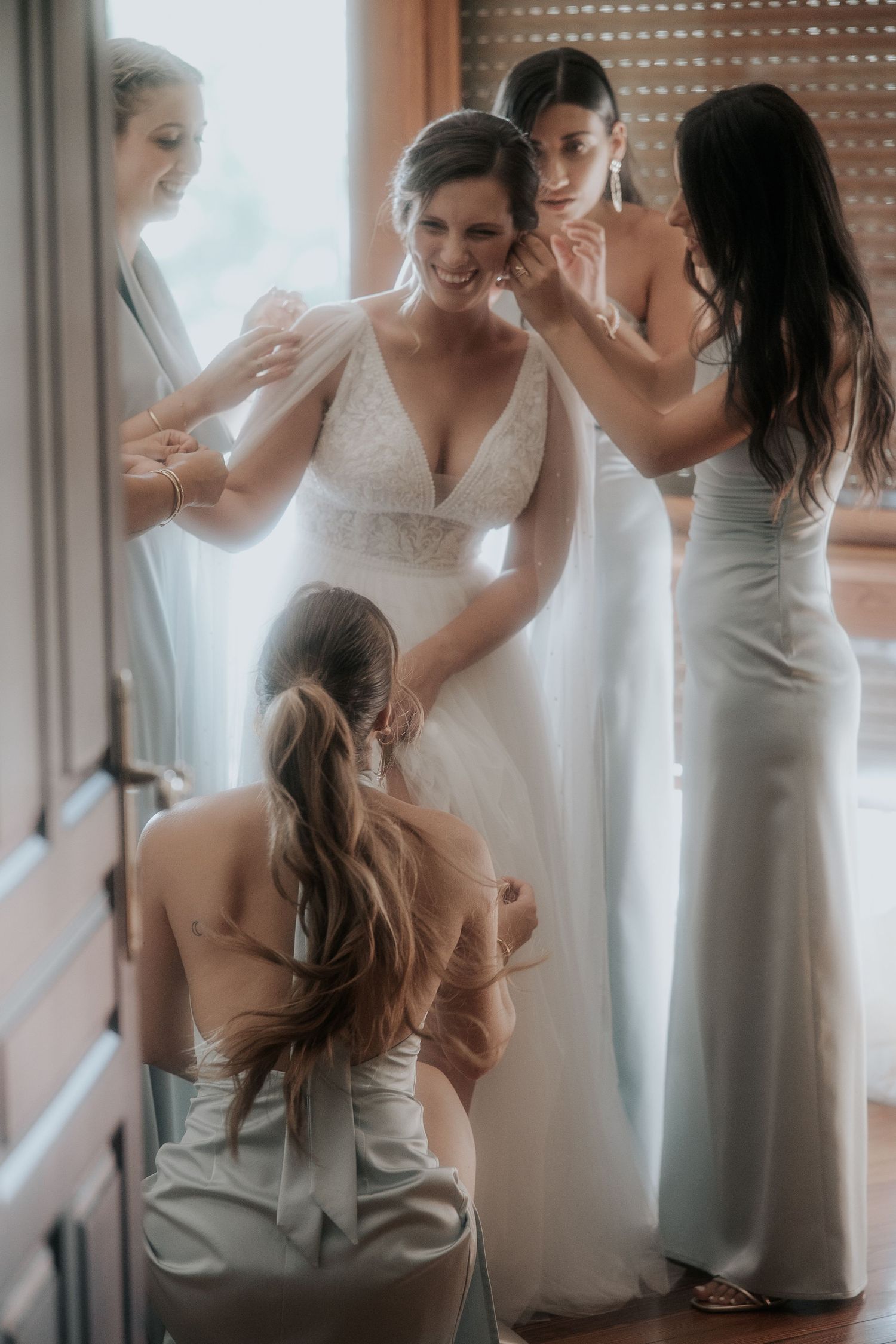 Wedding party assists with final dress preparations in natural window light.