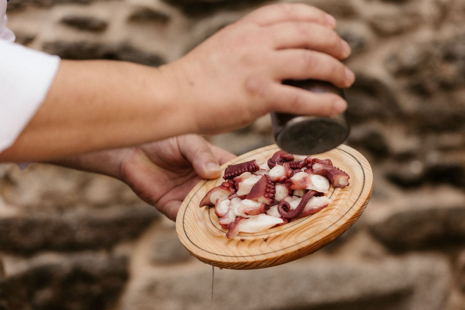 Hands placing sliced strawberries and whipped cream on a wooden plate in a rustic outdoor setting.