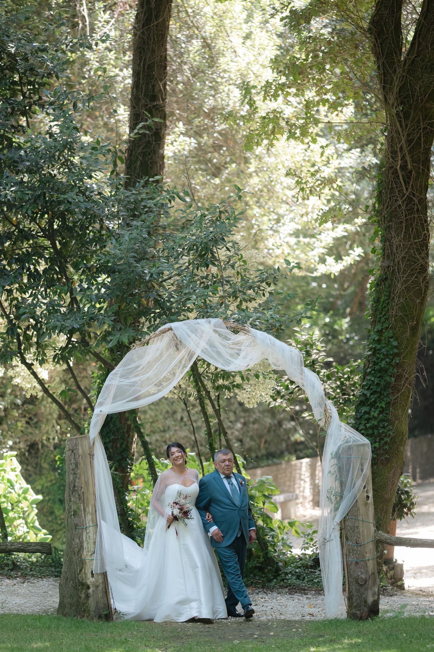 A romantic wedding scene under a white draped arch in a garden setting with trees and natural lighting.