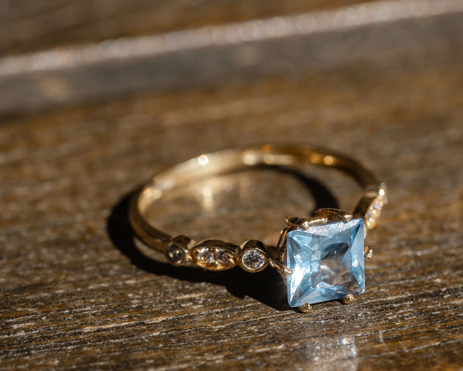 A delicate gold ring featuring a square-cut light blue topaz gemstone on a textured band rests on wooden surface.