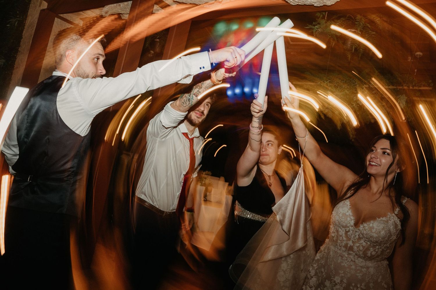 Wedding guests dancing and celebrating under warm lighting in a rustic venue with wooden beams overhead.