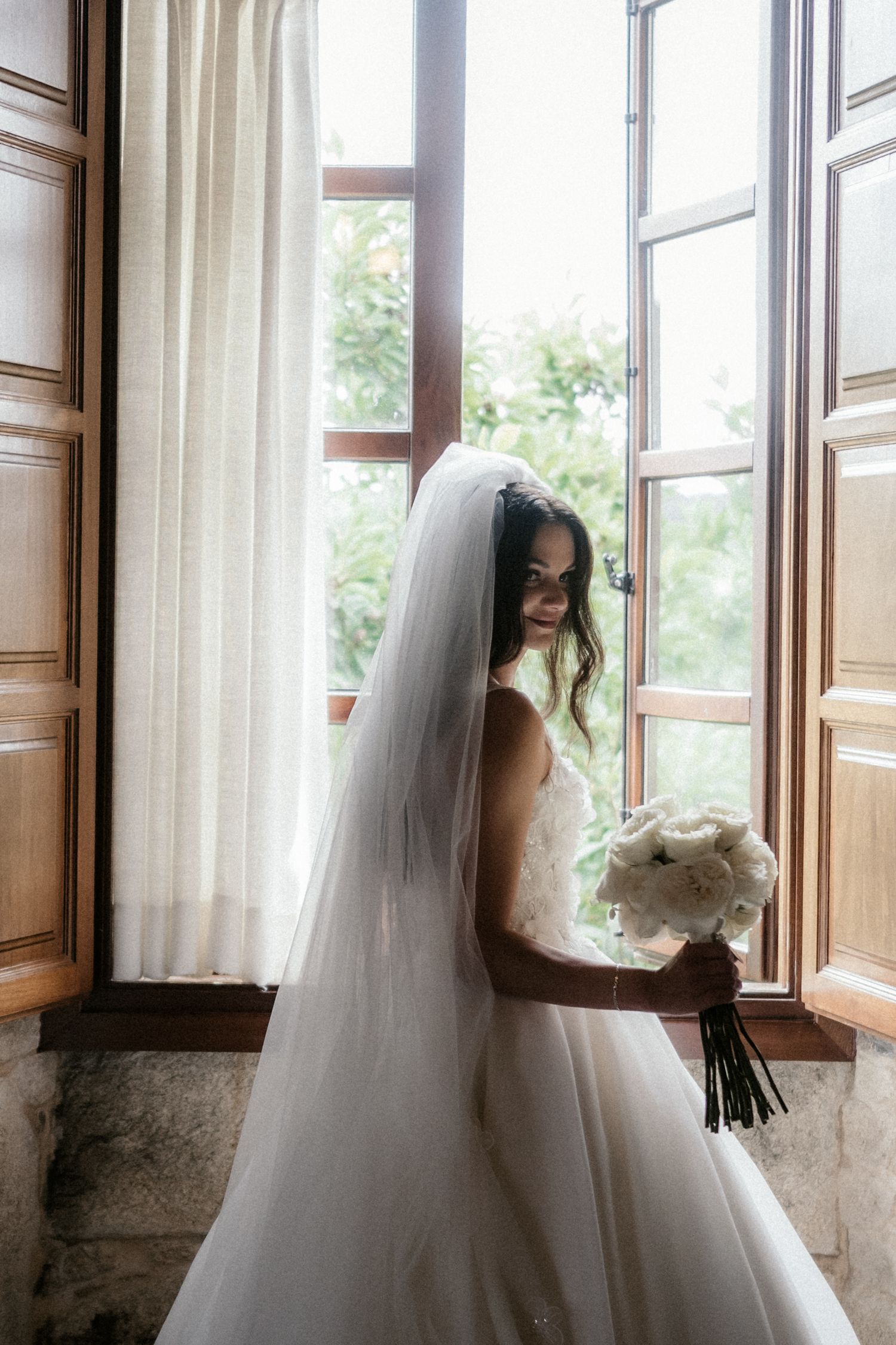 A bride in a white wedding gown gazes out multiple windows in a series of romantic bridal portraits.