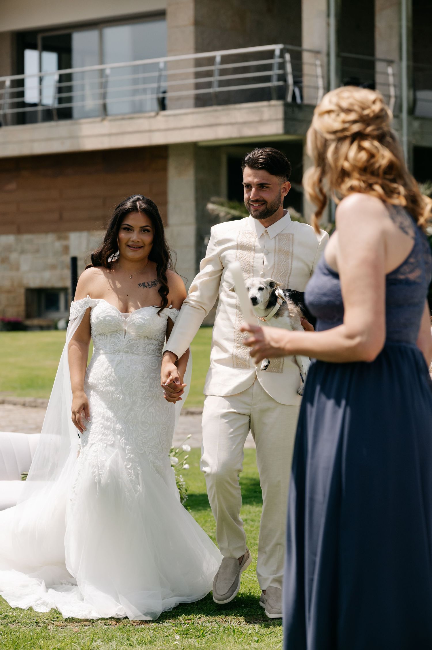 A wedding party in navy blue and white dresses poses outdoors on a sunny day with a small white dog.