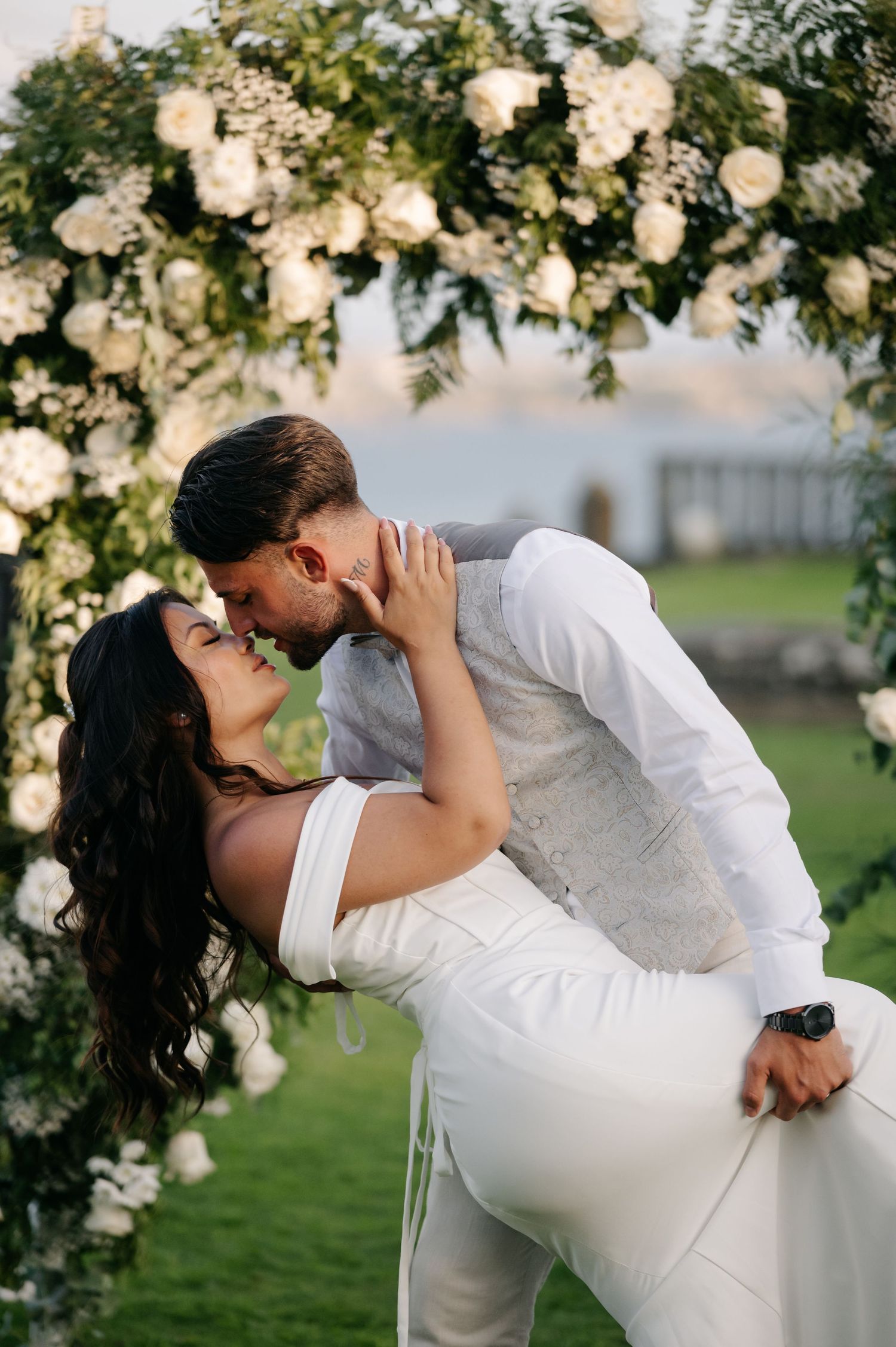 Romantic wedding couple shares passionate kisses under floral arch decorations in outdoor garden setting.