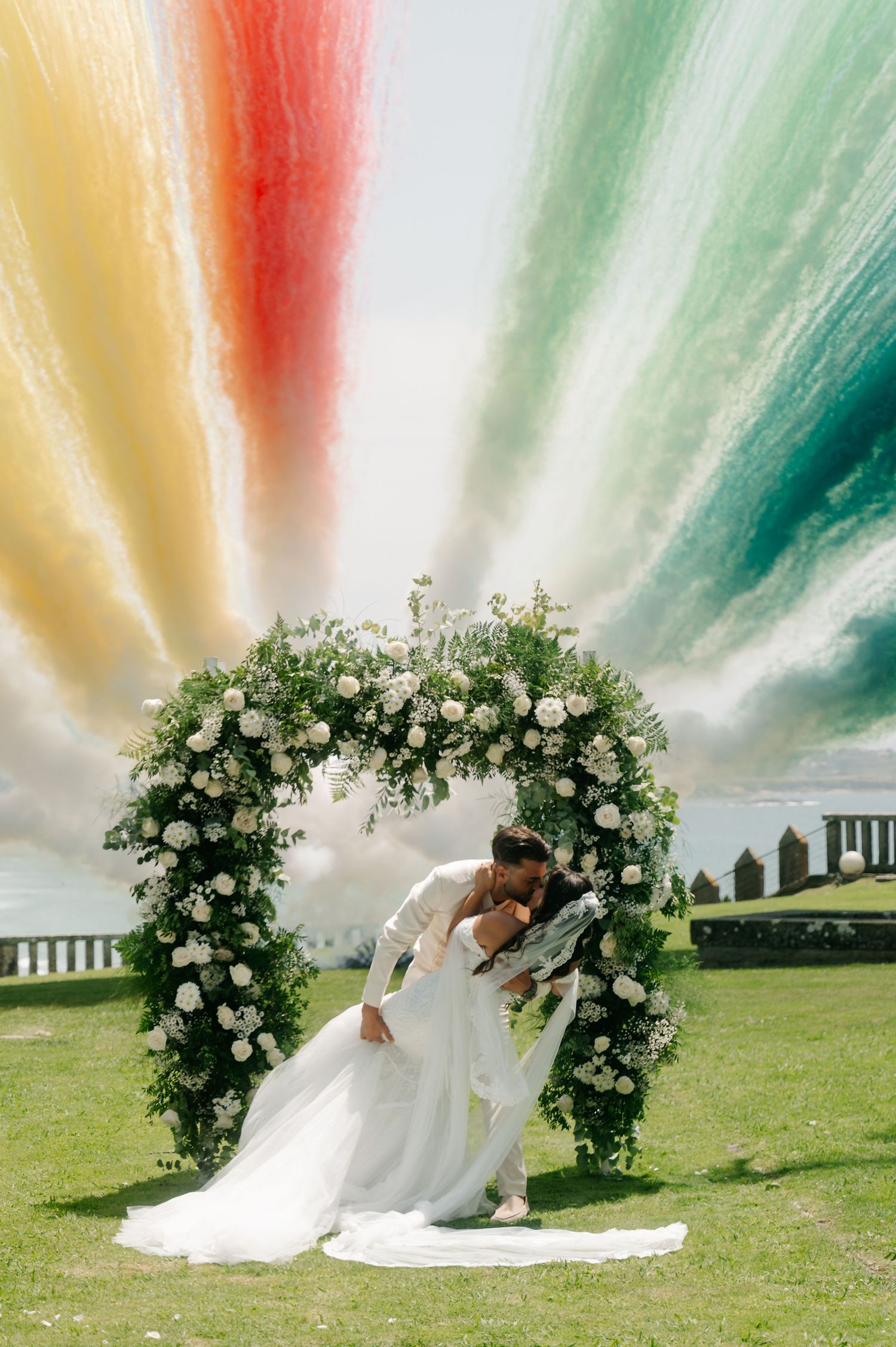 A romantic wedding couple dips under a floral arch in a grassy outdoor setting with colorful streaks in the sky.
