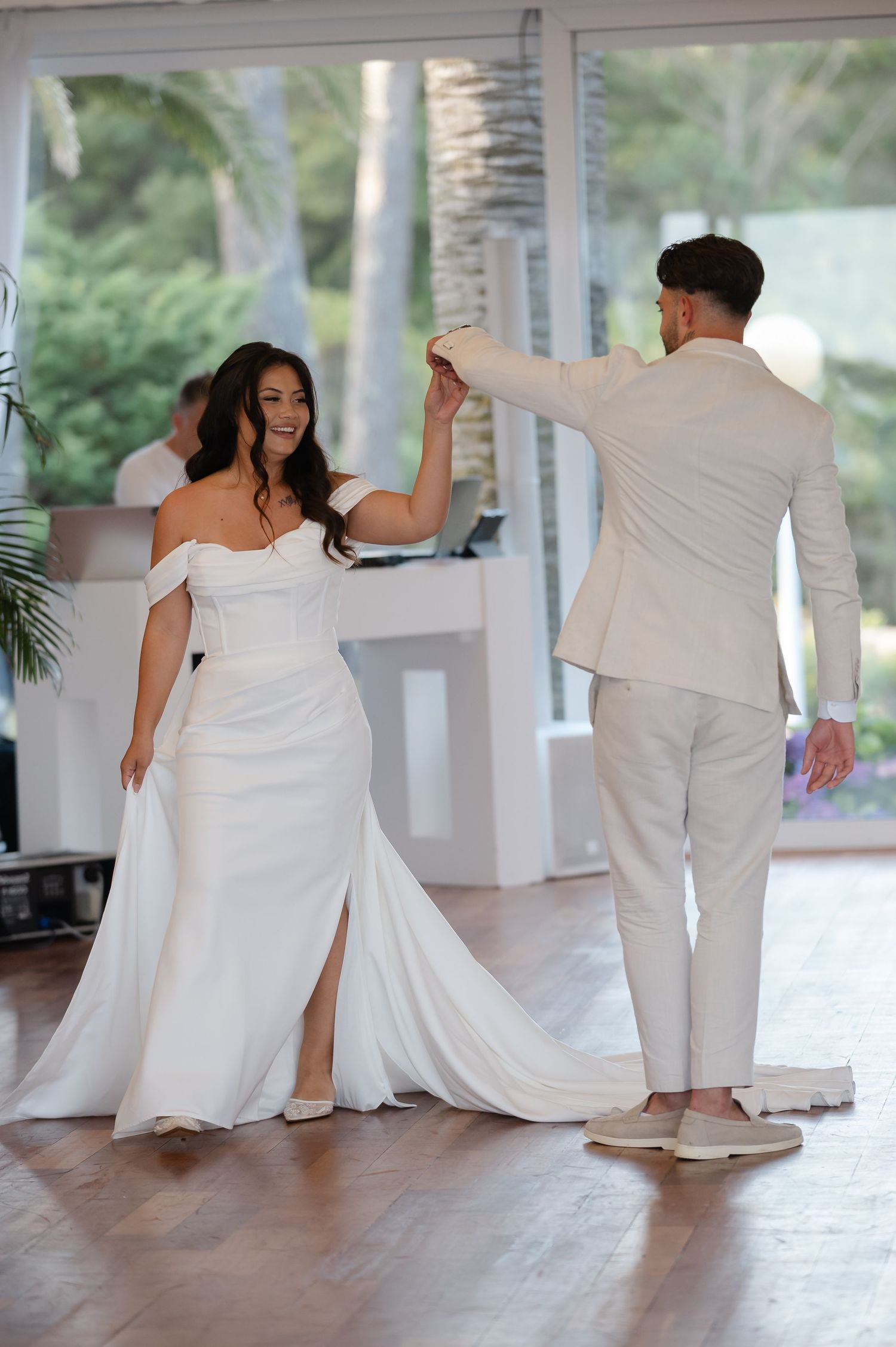 A couple shares a joyful dance moment at their wedding, with the bride in a flowing white gown and groom in light beige suit.