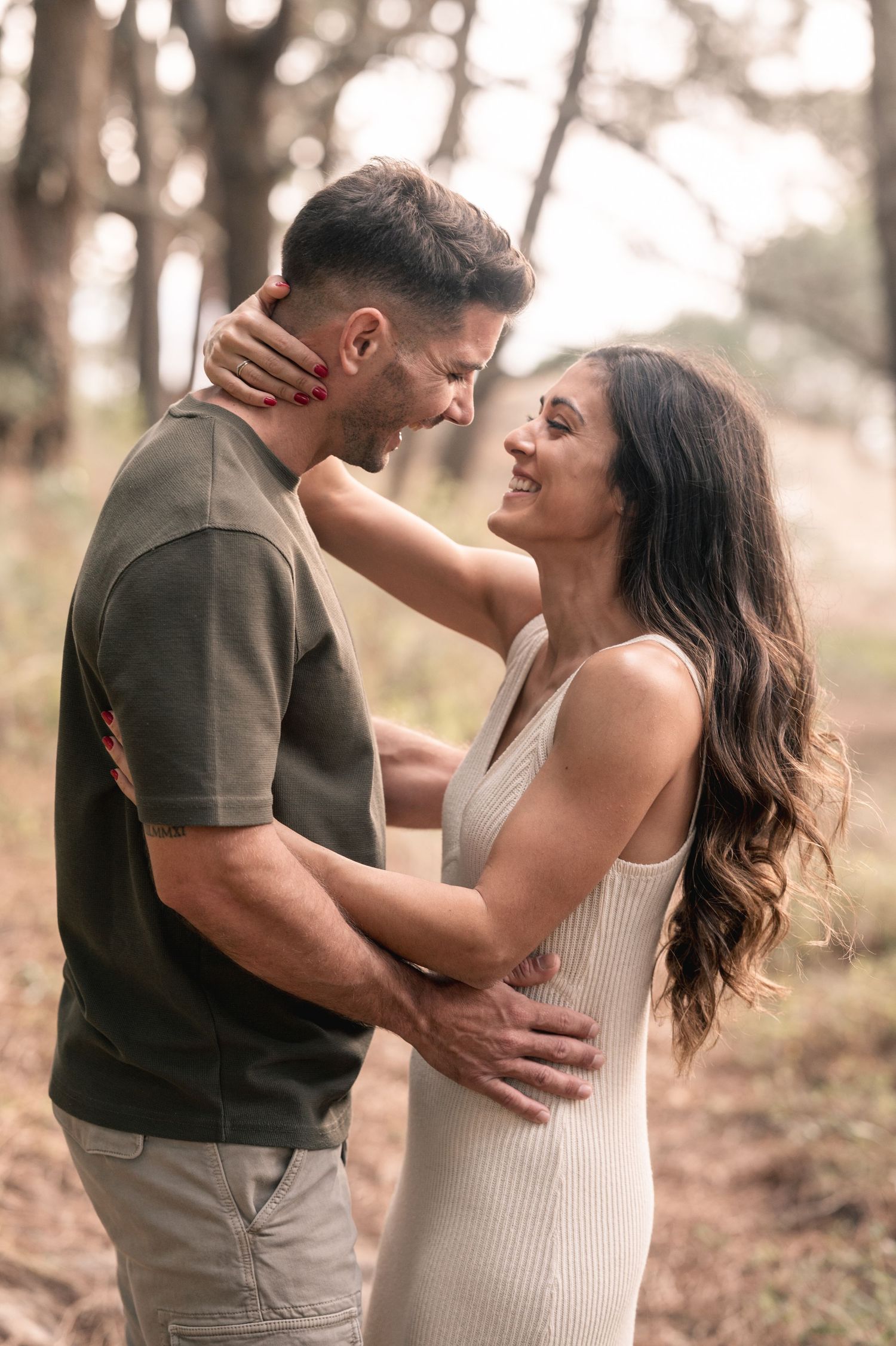A romantic couple embraces and laughs together in a sun-dappled forest setting wearing casual clothing.