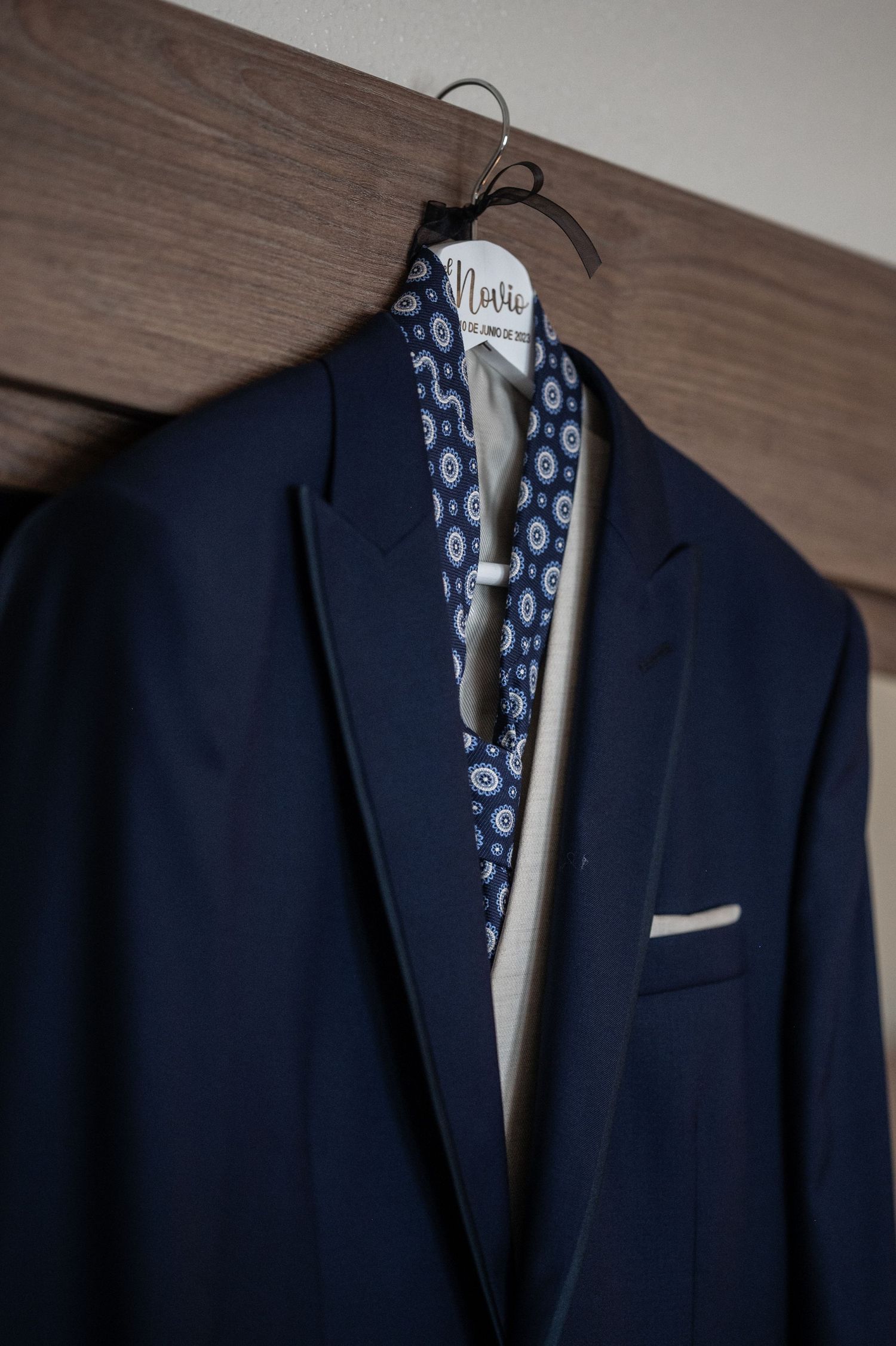 A navy blue suit jacket hangs on a wooden hanger against a textured wall with patterned fabric detail.