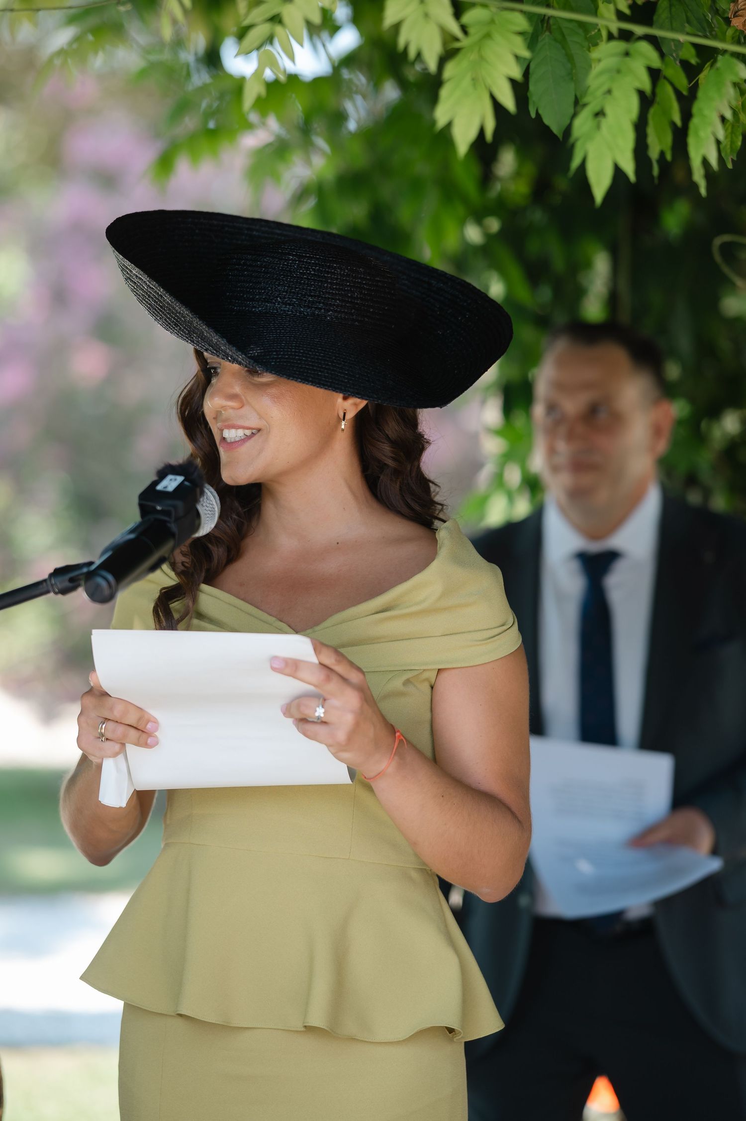 Person in a wide-brimmed black hat and beige dress smiles while holding papers during an outdoor event.