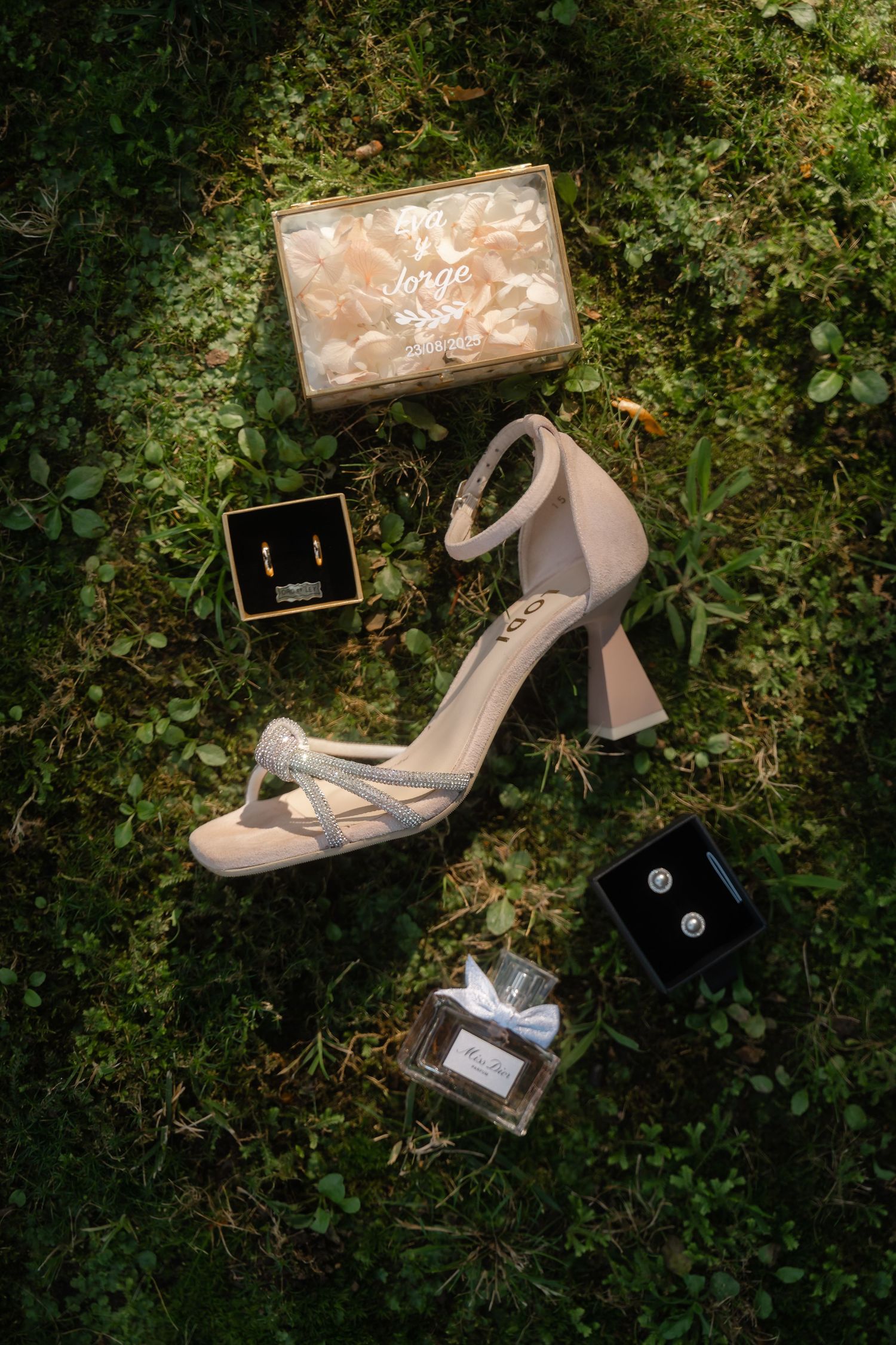 Elegant silver bridal shoes and accessories arranged on green grass with jewelry boxes.