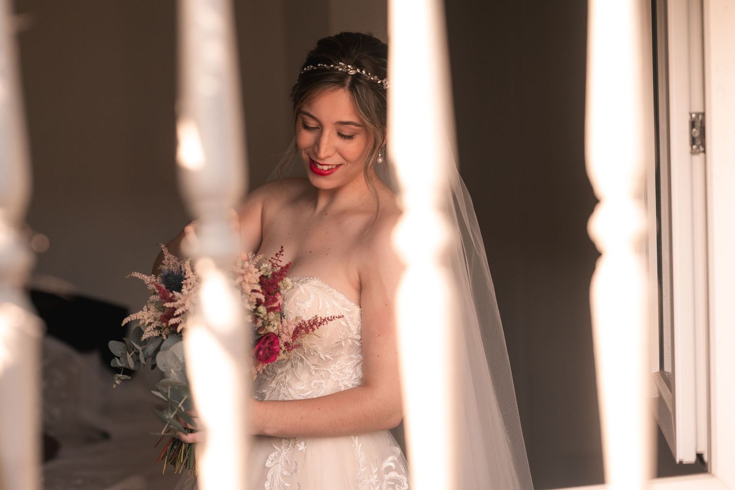 A bride in a white lace wedding dress poses by a window with soft natural light streaming in.