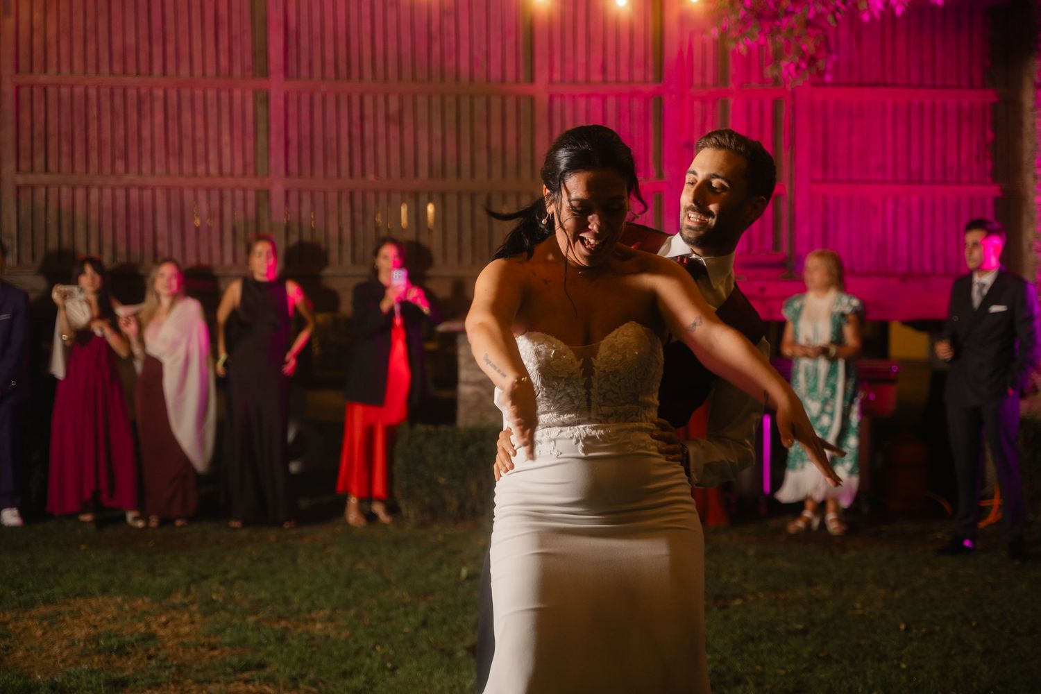 Wedding guests dance on an outdoor dance floor illuminated by string lights and pink uplighting at night.