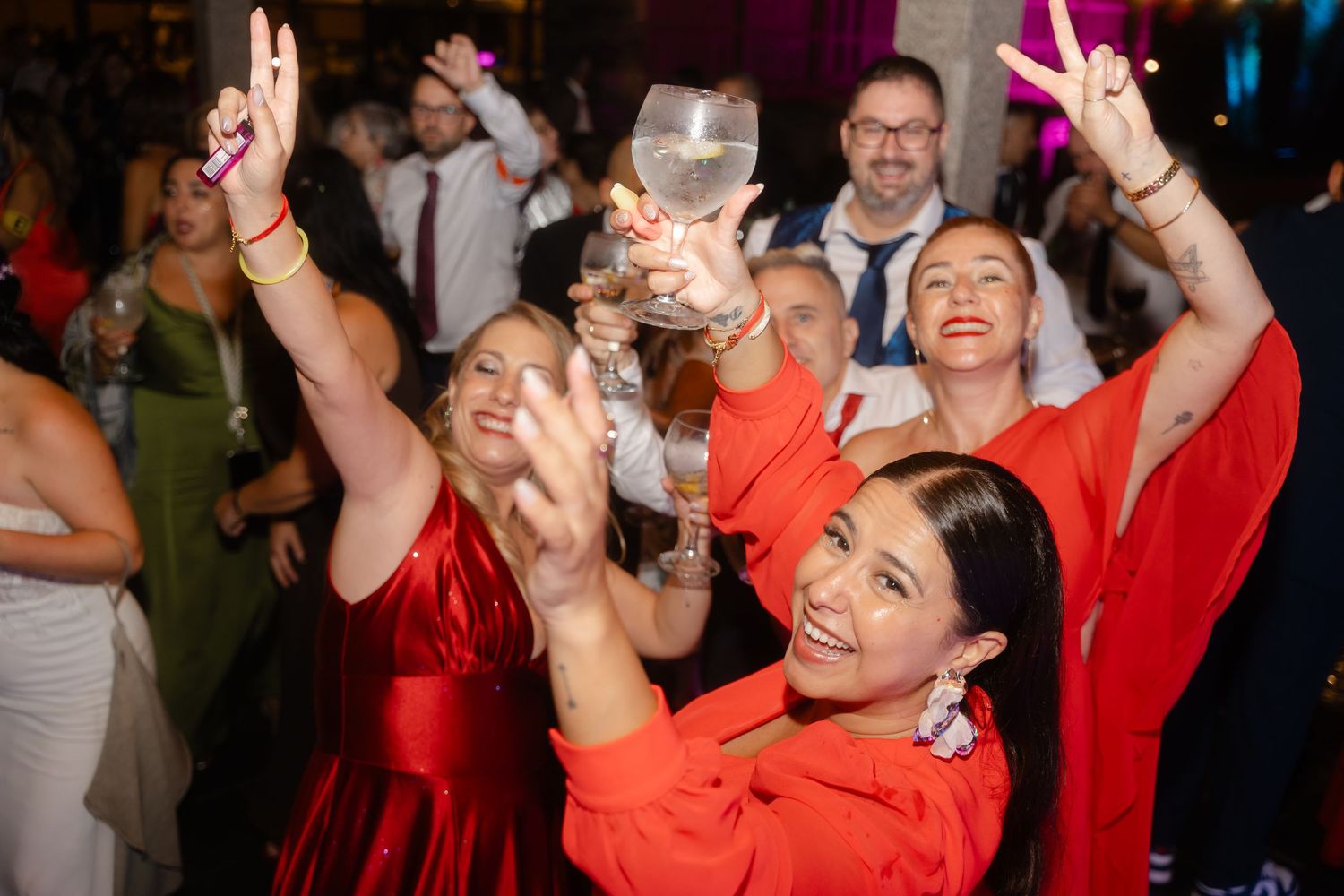Energetic wedding guests dancing and celebrating with hands raised under dramatic party lighting.
