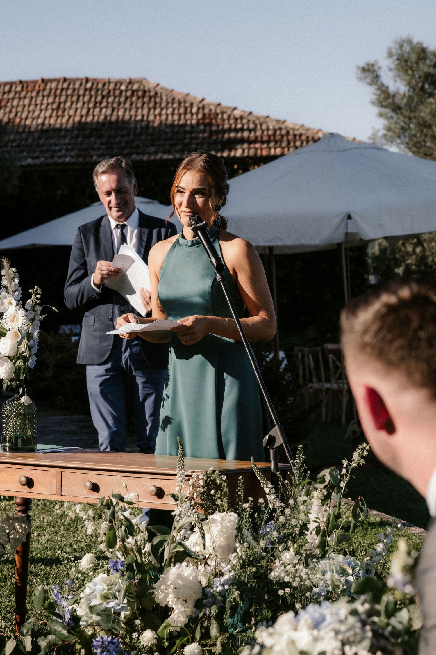 Someone in a sage green dress speaks at a podium decorated with white floral arrangements during an outdoor ceremony.