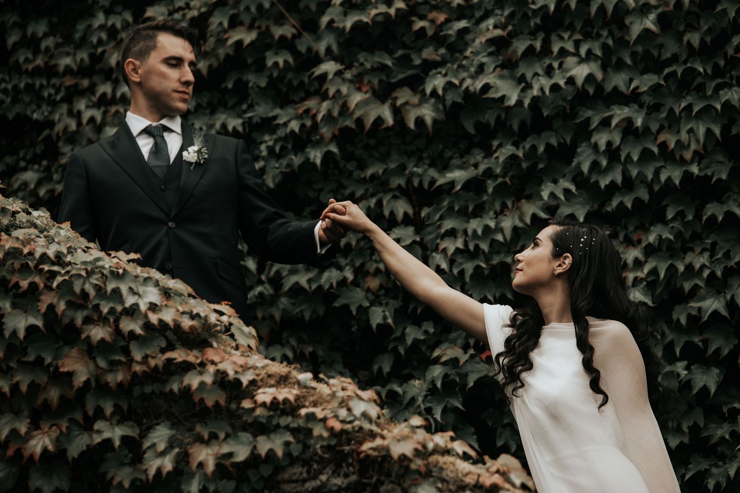 A romantic wedding couple embraces in front of a lush ivy-covered wall in moody natural lighting.