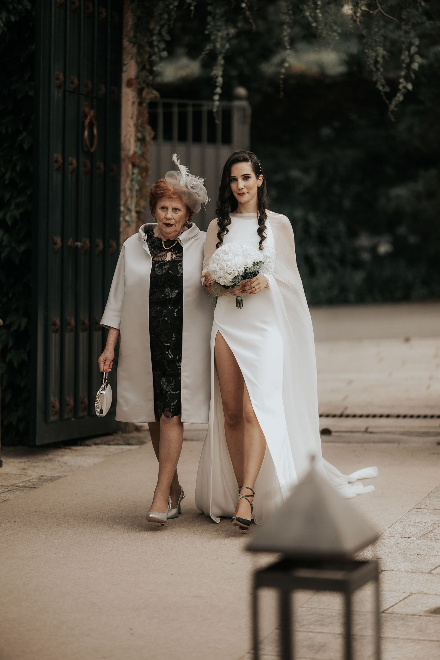 Two people in contrasting black and white outfits stand together in an outdoor setting with wrought iron gates.