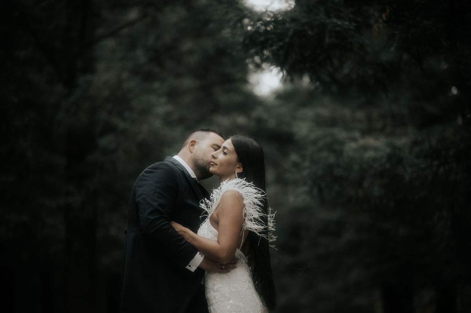 A romantic wedding couple shares an intimate kiss in a dark wooded setting with dramatic lighting and shadows.