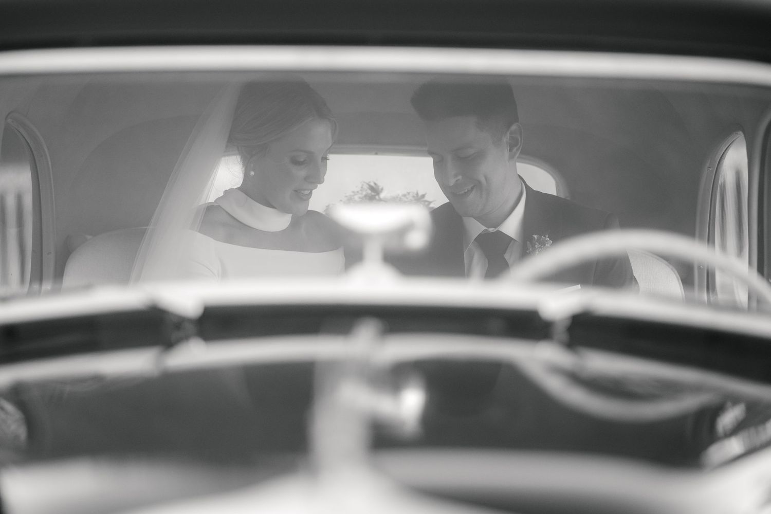A black and white vintage photograph shows two people sitting together in the front seat of a classic car.