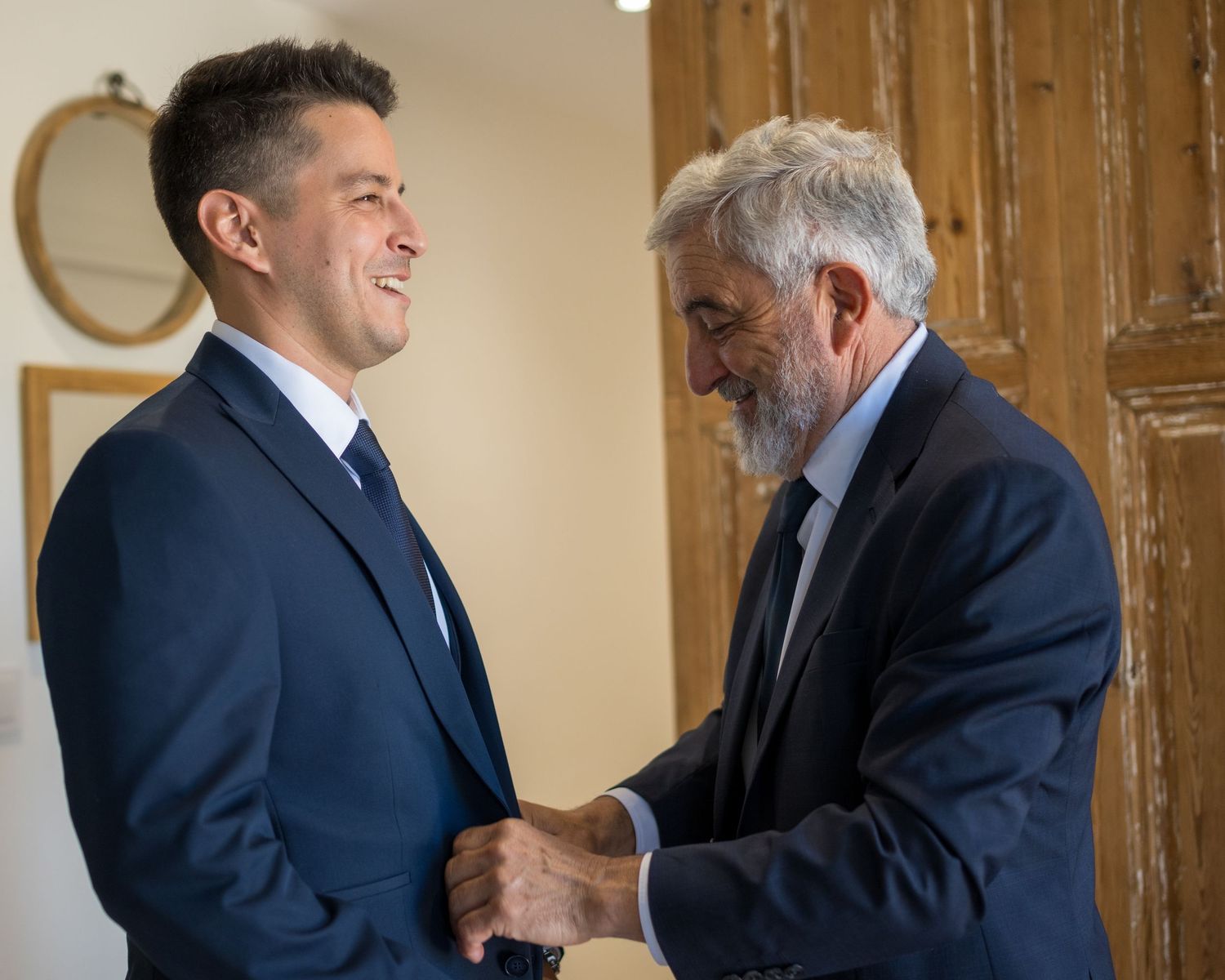 Two people in business suits shake hands in a professional setting with wooden panels in the background.