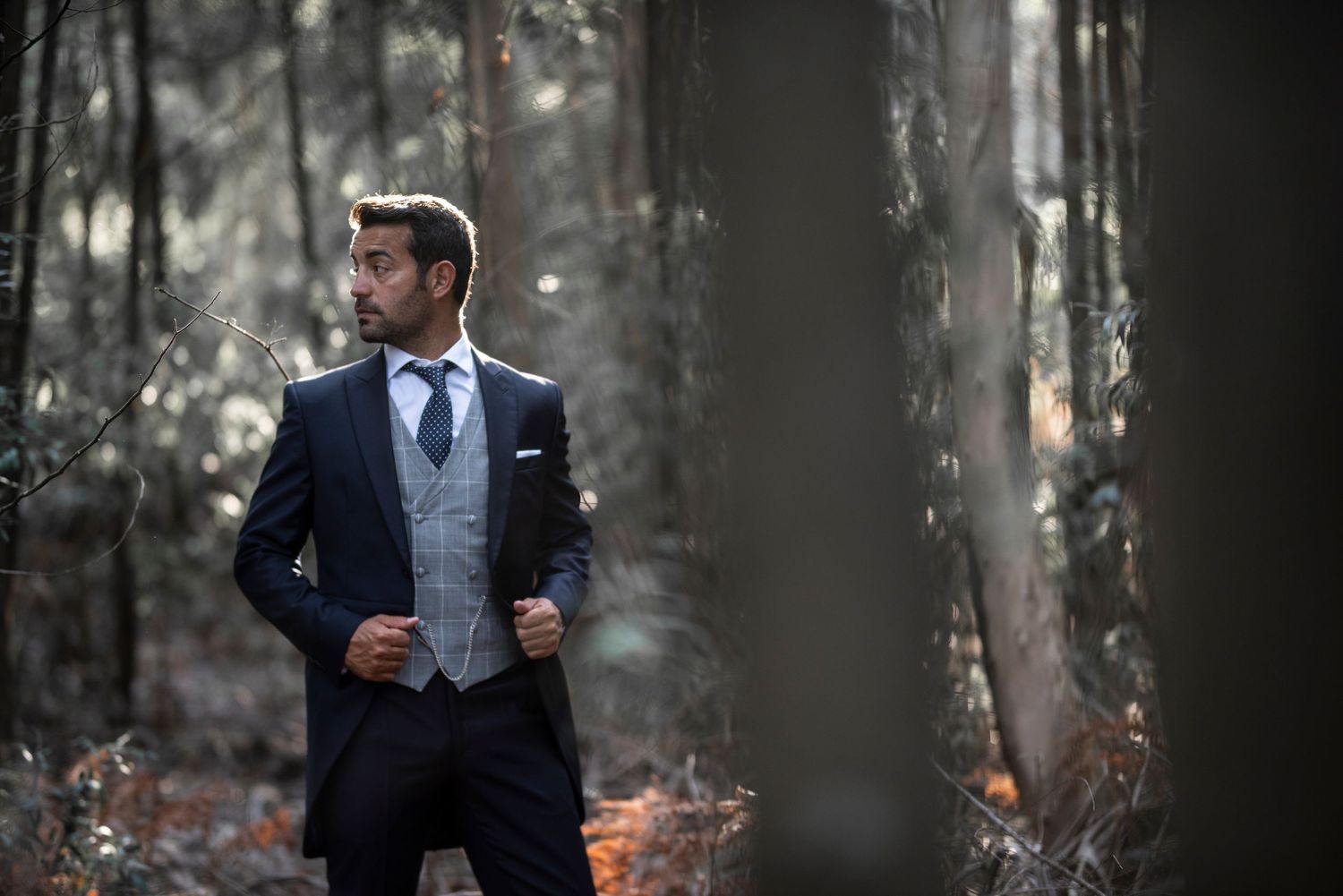 A well-dressed man in a navy suit walks through a misty autumn forest holding papers and a coffee cup.
