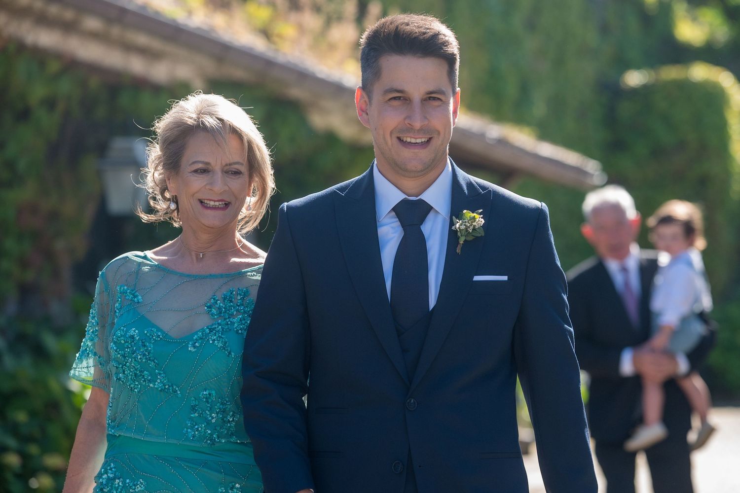 Wedding guests in formal attire walk together on a sunny day through an outdoor venue wearing blue and teal outfits.