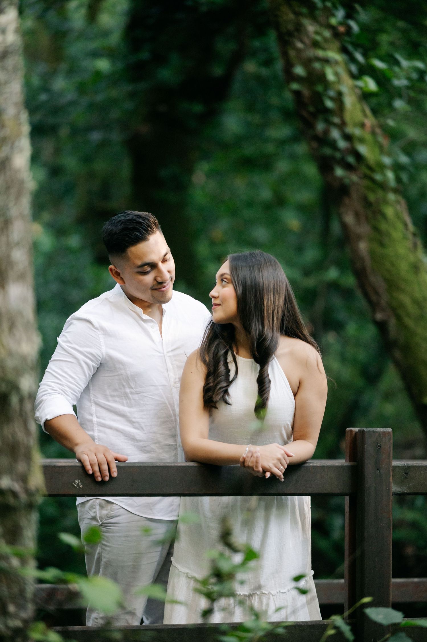 A couple in coordinated white outfits share a loving moment while leaning against a wooden railing in a forest.