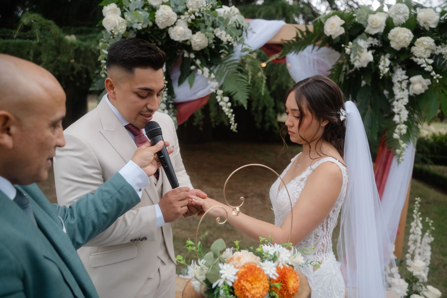 A wedding ceremony under a floral arch with white roses as the couple exchanges rings during their vows.