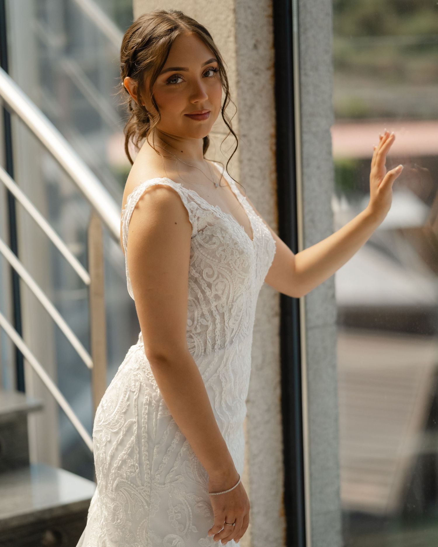 A bride in a white lace wedding dress poses elegantly against a window with modern railings.