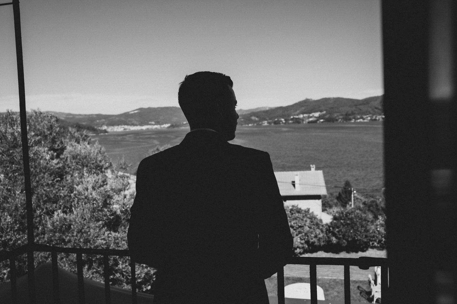 A silhouetted figure stands on a balcony overlooking a coastal Mediterranean landscape with mountains and sea views.