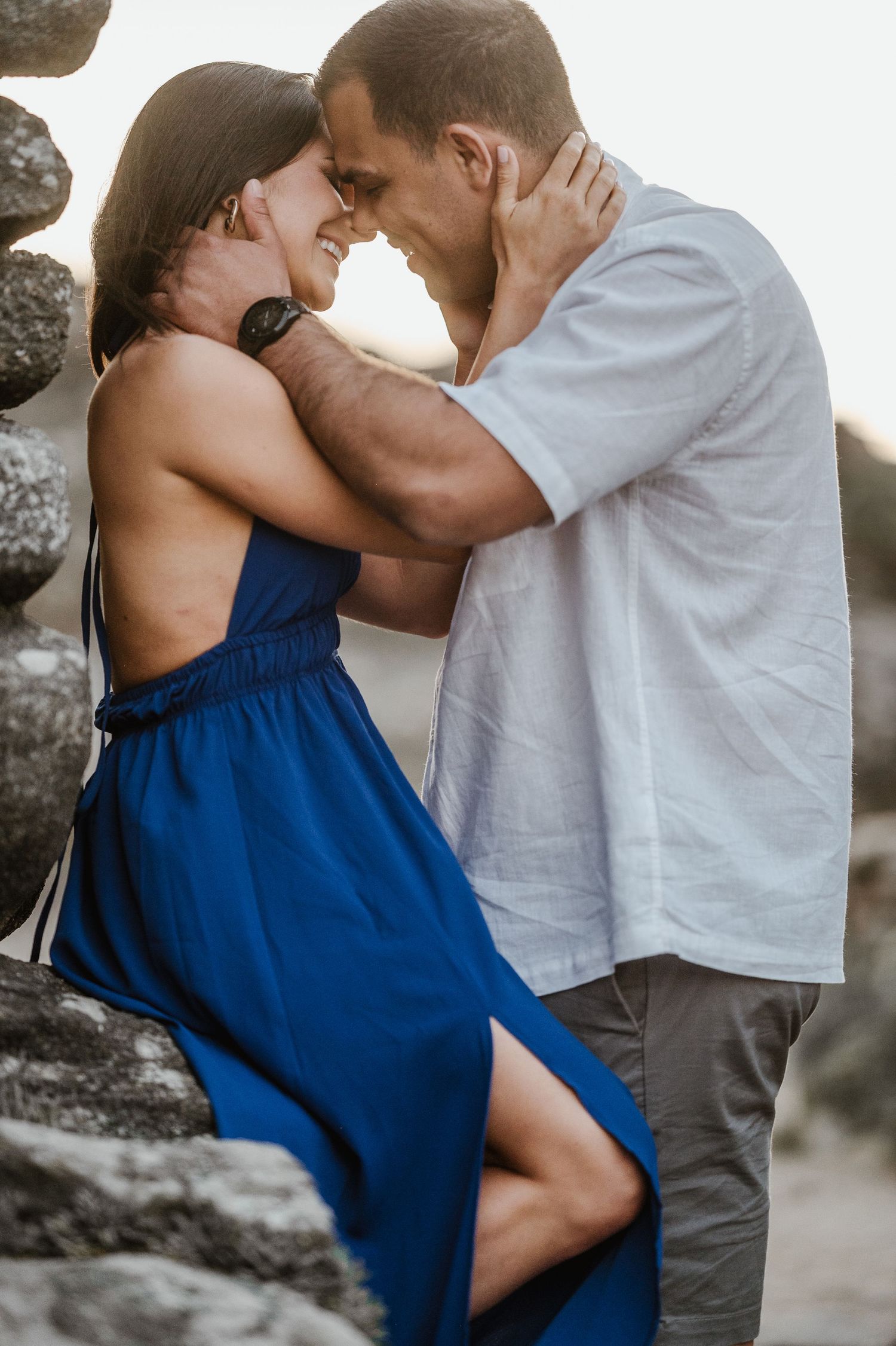 Romantic couple in blue dress and white shirt sharing intimate moments against stone wall at sunset.
