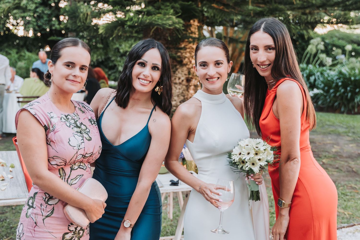 Bridesmaids in coral and teal dresses pose together outdoors at a summer wedding celebration.