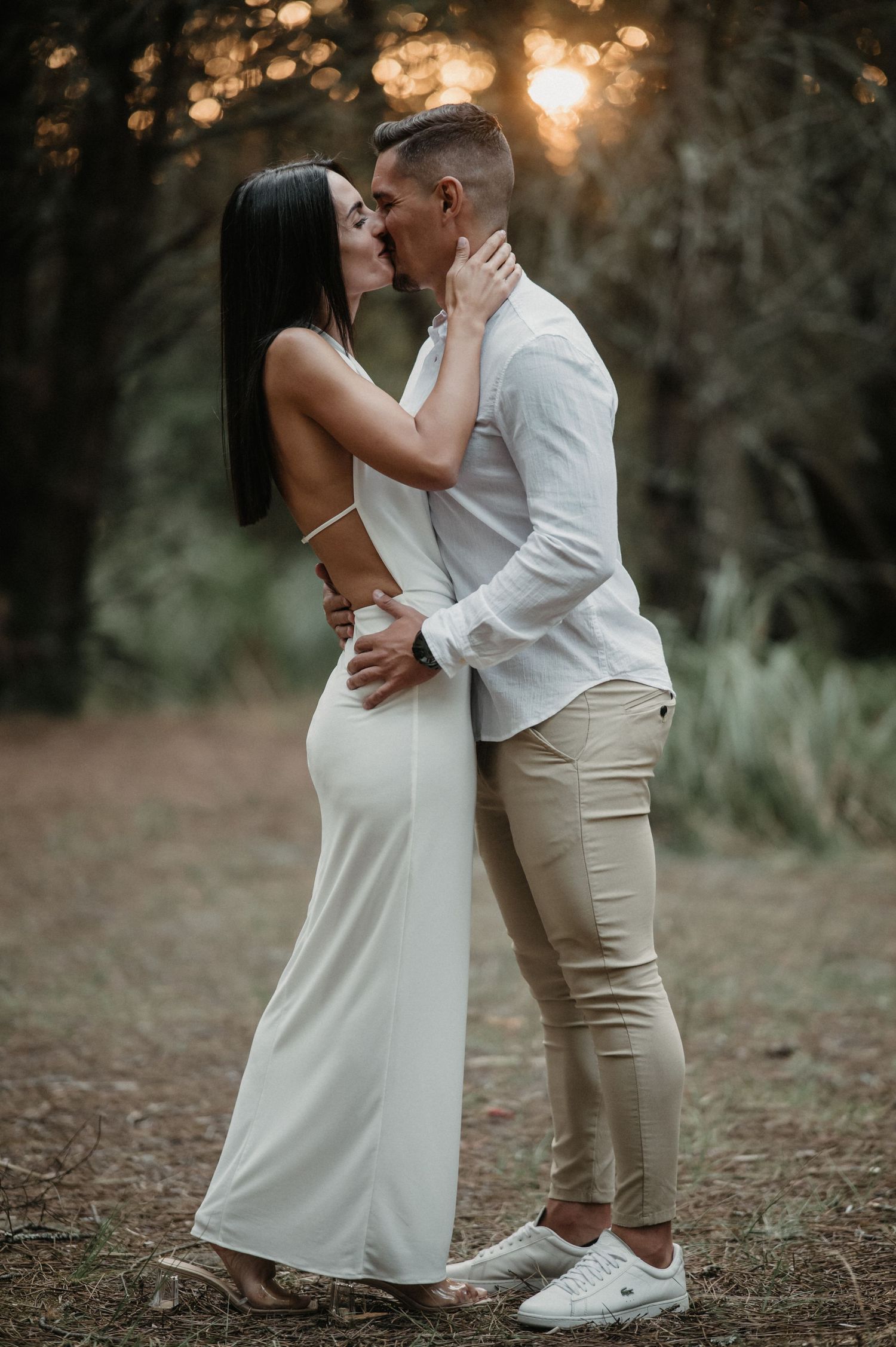 Intimate moment between couple in coordinated white and beige outfits against sunset forest backdrop.