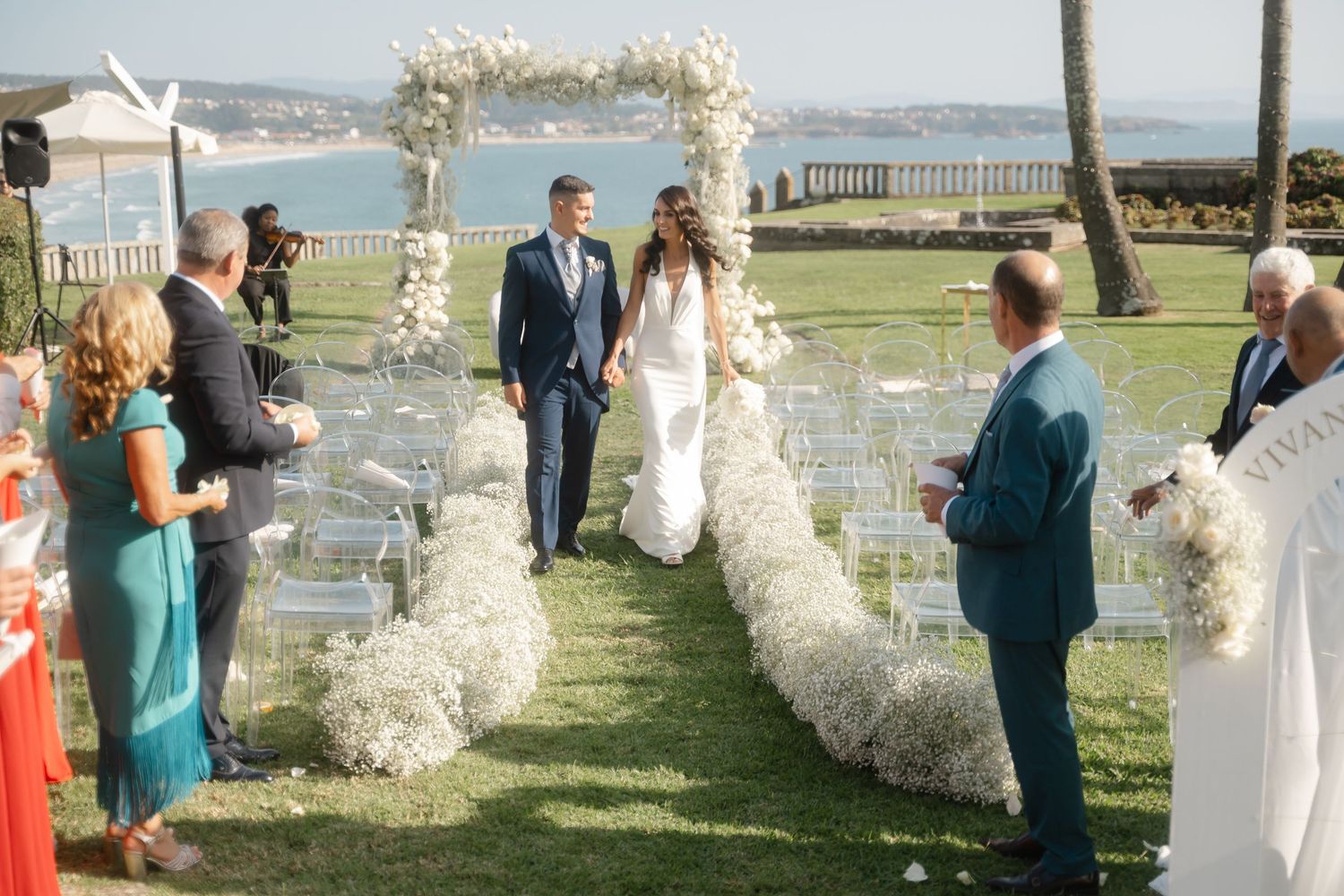 Wedding guests gather on a grassy lawn with ocean views for an outdoor ceremony under floral arches.