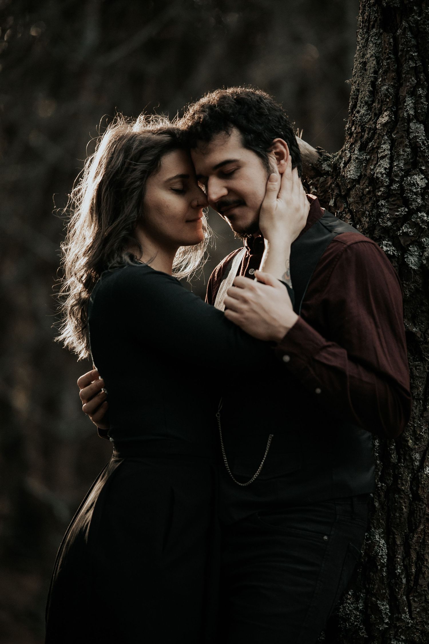 A romantic couple in dark clothing share an intimate moment against a tree in moody lighting.