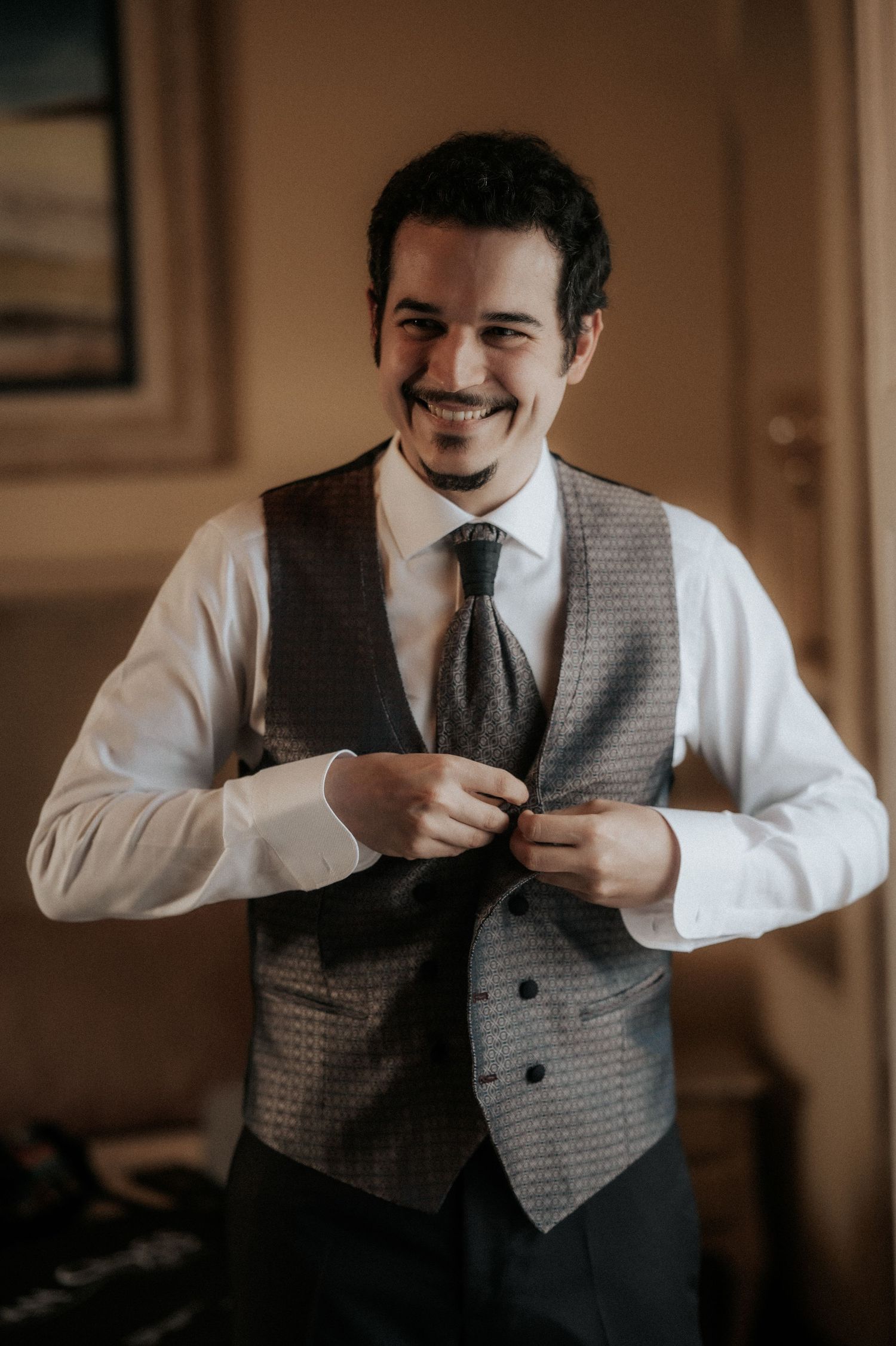 A groom in formal attire buttons up his textured grey vest while getting ready, smiling warmly in warm indoor lighting.