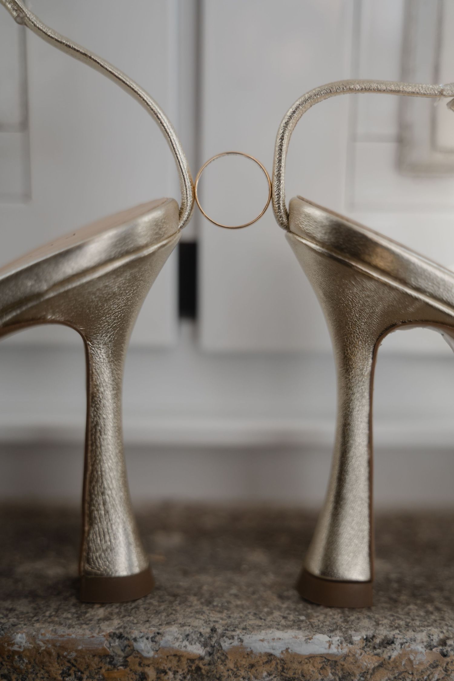 Close-up of metallic gold wedding shoes with stiletto heels and a wedding ring balanced between them.