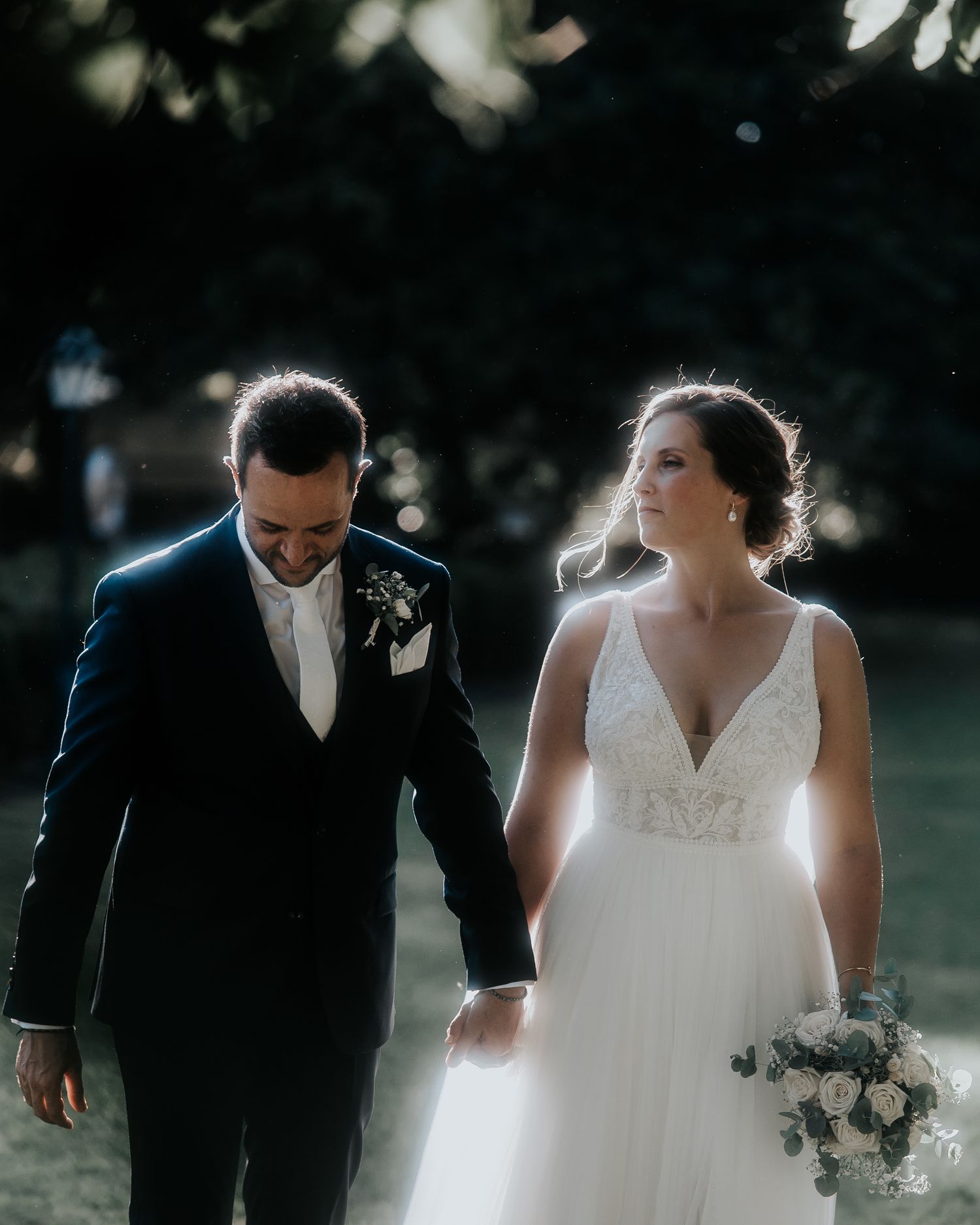 A romantic wedding couple walks together outdoors in a moody, cinematic scene with natural lighting.