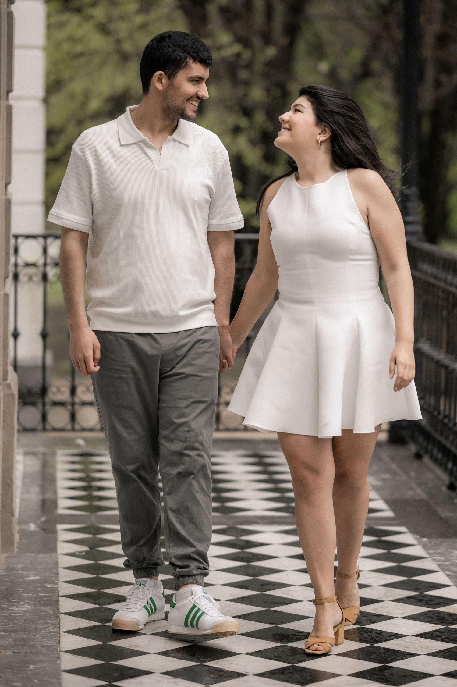 Couple in coordinated white outfits walk playfully on a checkered floor walkway.