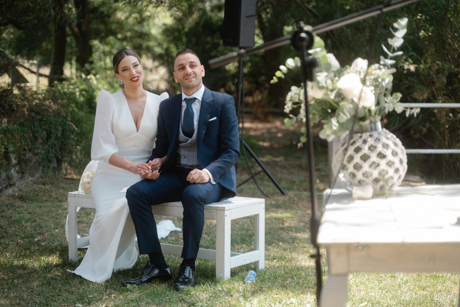Wedding ceremony seating arrangement with white benches set up outdoors near trees.