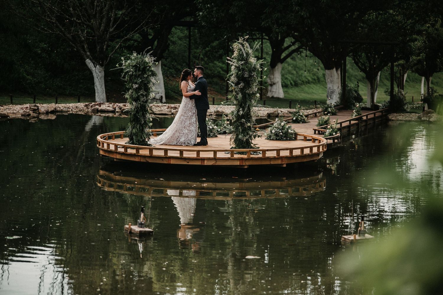 Couple in formal attire poses for romantic wedding photos near a tranquil pond in a lush garden setting.