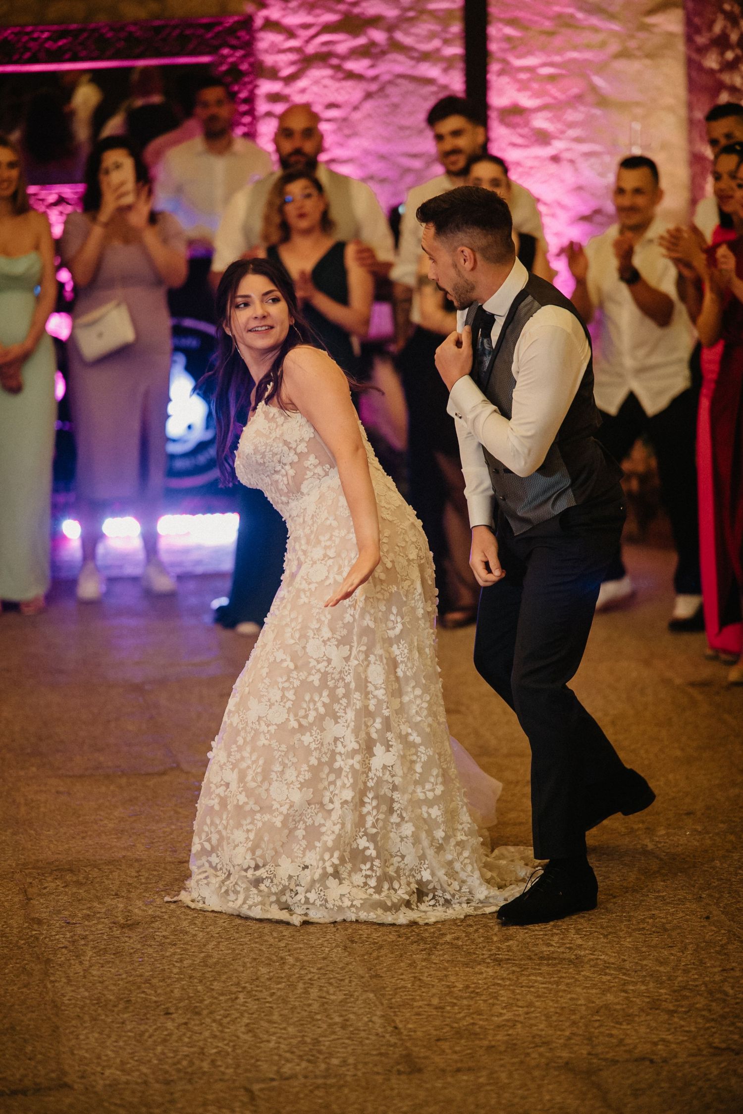 A couple dances together at a wedding reception with pink uplighting illuminating stone walls in the background.