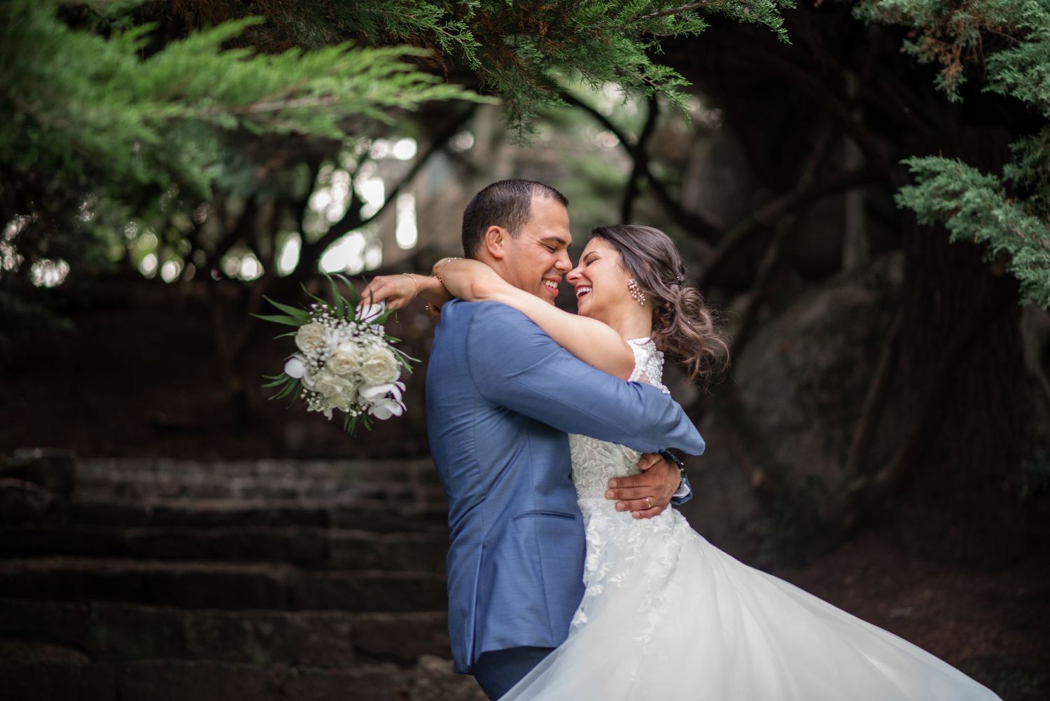 A couple embraces during their wedding ceremony in a lush garden setting with a white bouquet in hand.