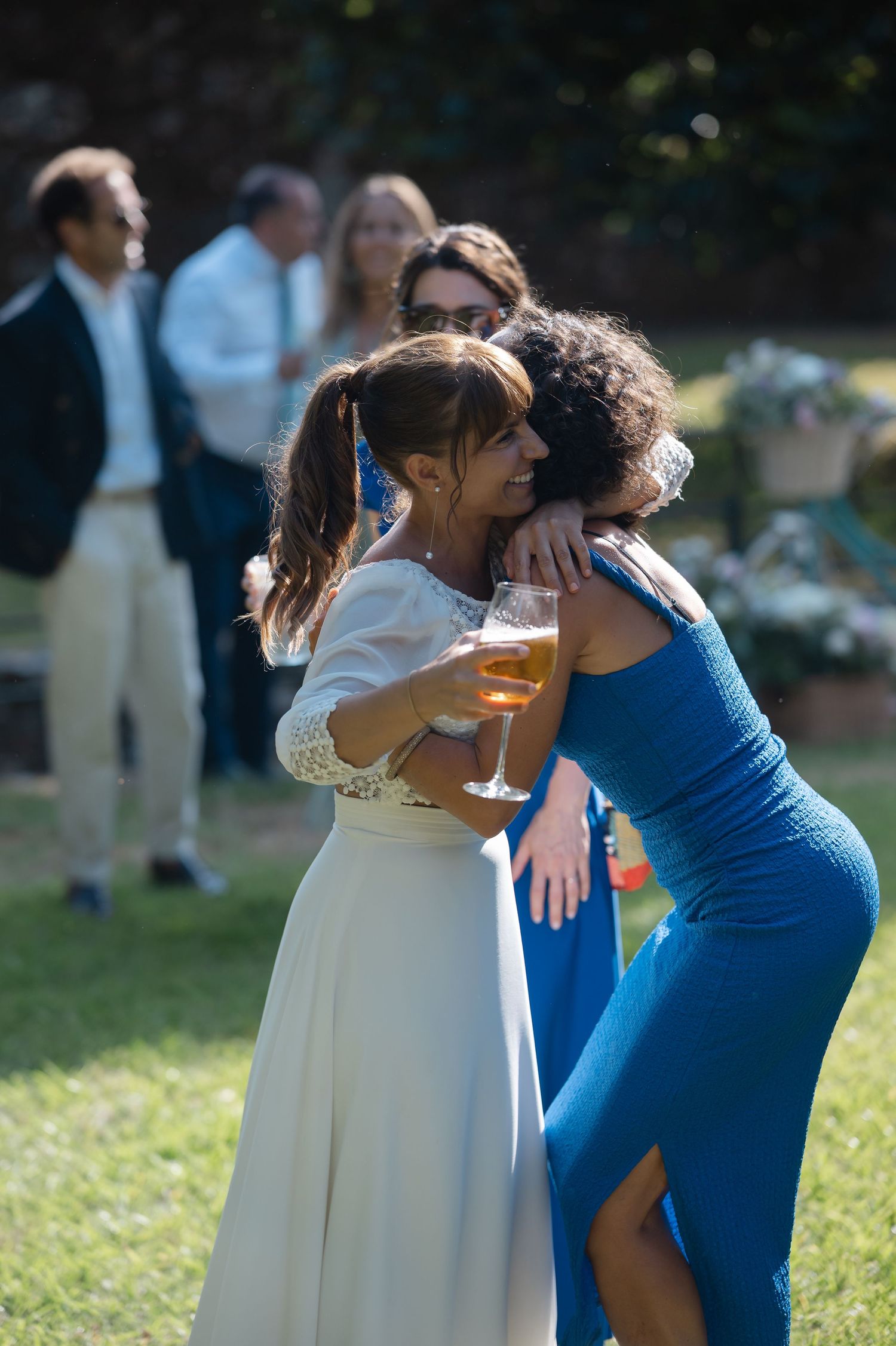 Wedding guests in white and blue dresses hugging and holding drinks during an outdoor celebration.