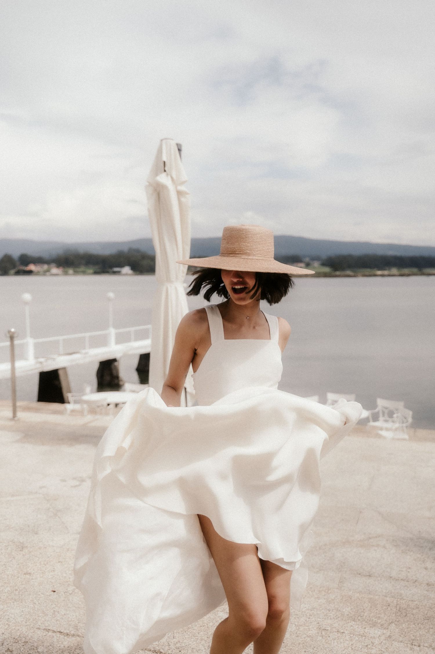 A flowing white dress billows in the ocean breeze as someone walks along a seaside railing on a cloudy day.
