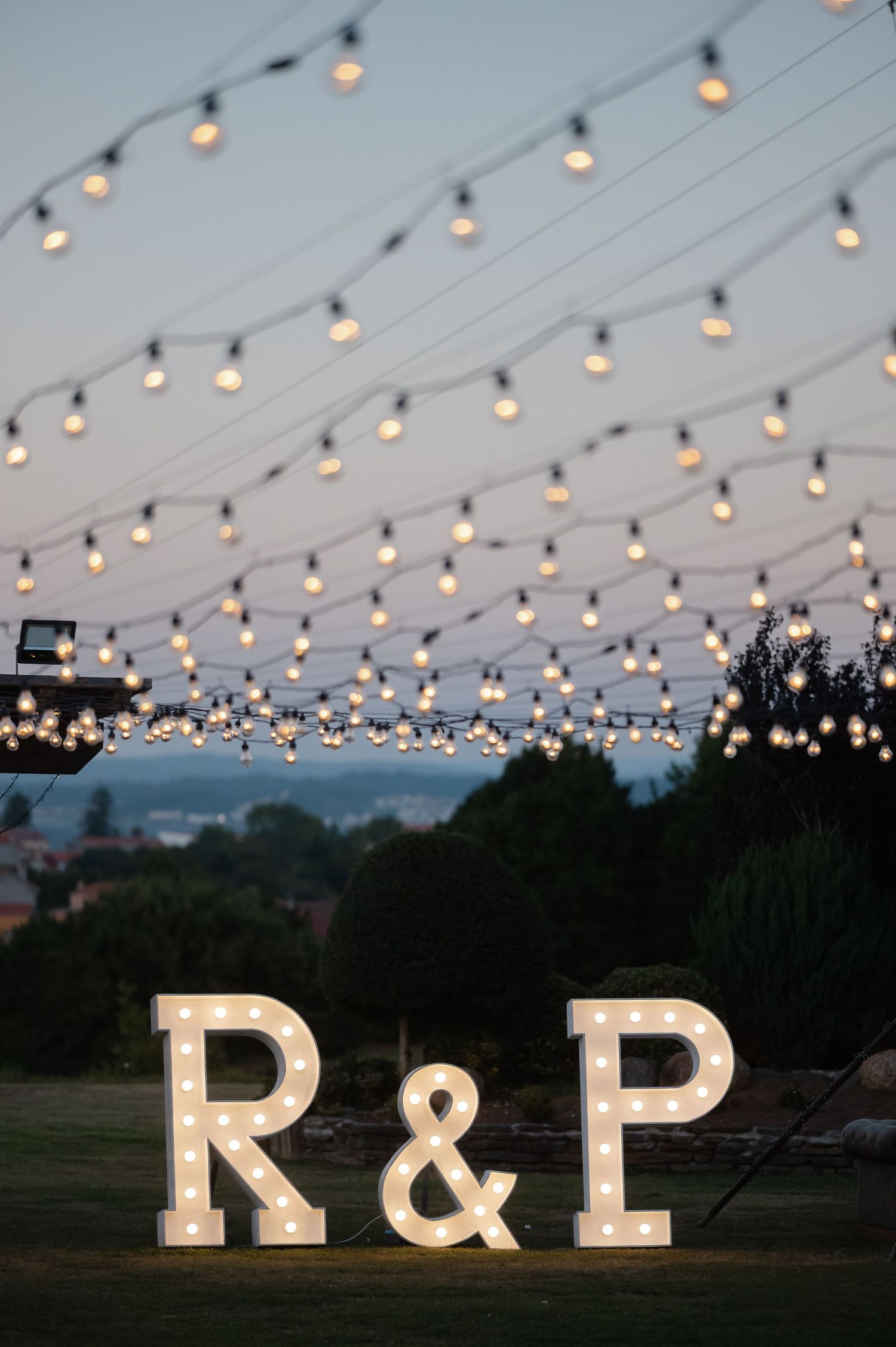 String lights illuminate an outdoor wedding reception with illuminated marquee letters R & P displayed at dusk.