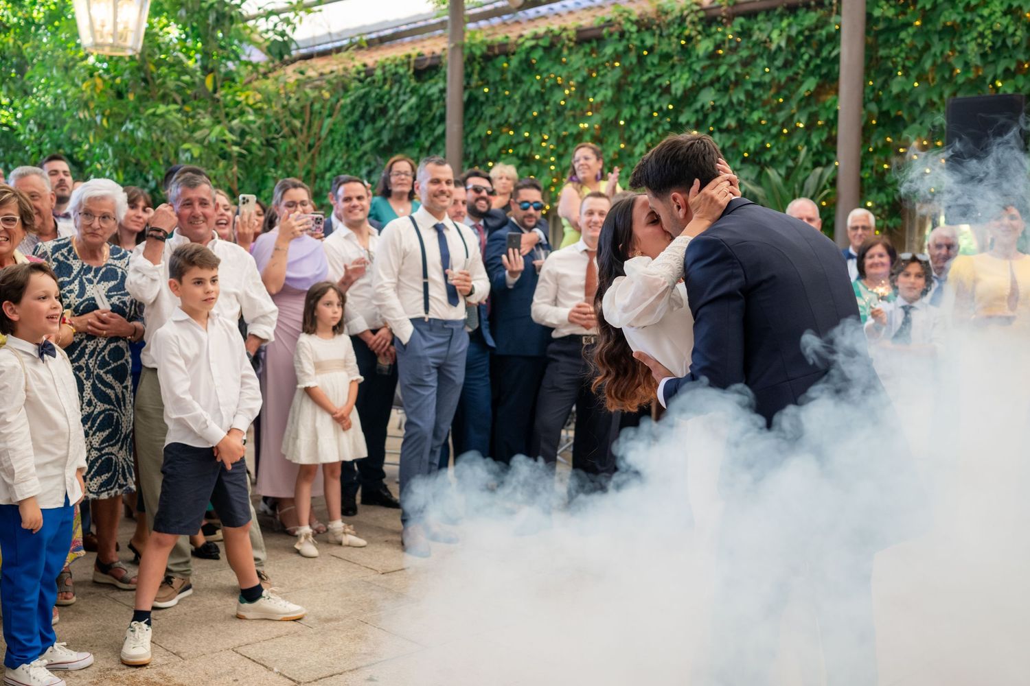Wedding celebration in an outdoor garden setting with guests gathered along a path lined with greenery.