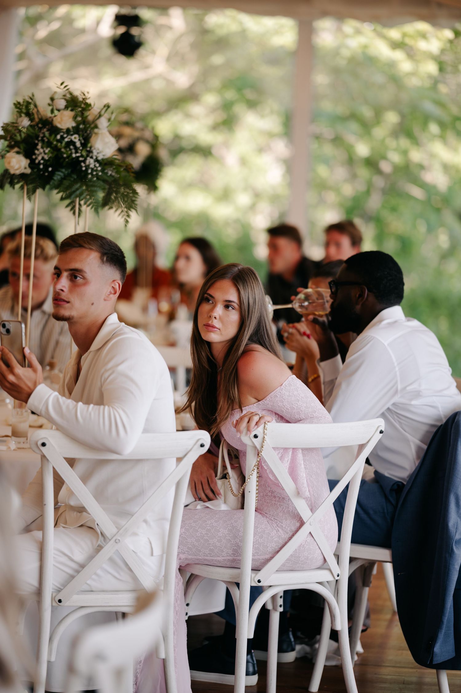 Wedding guests seated at white chairs under a tent during an outdoor reception celebration.