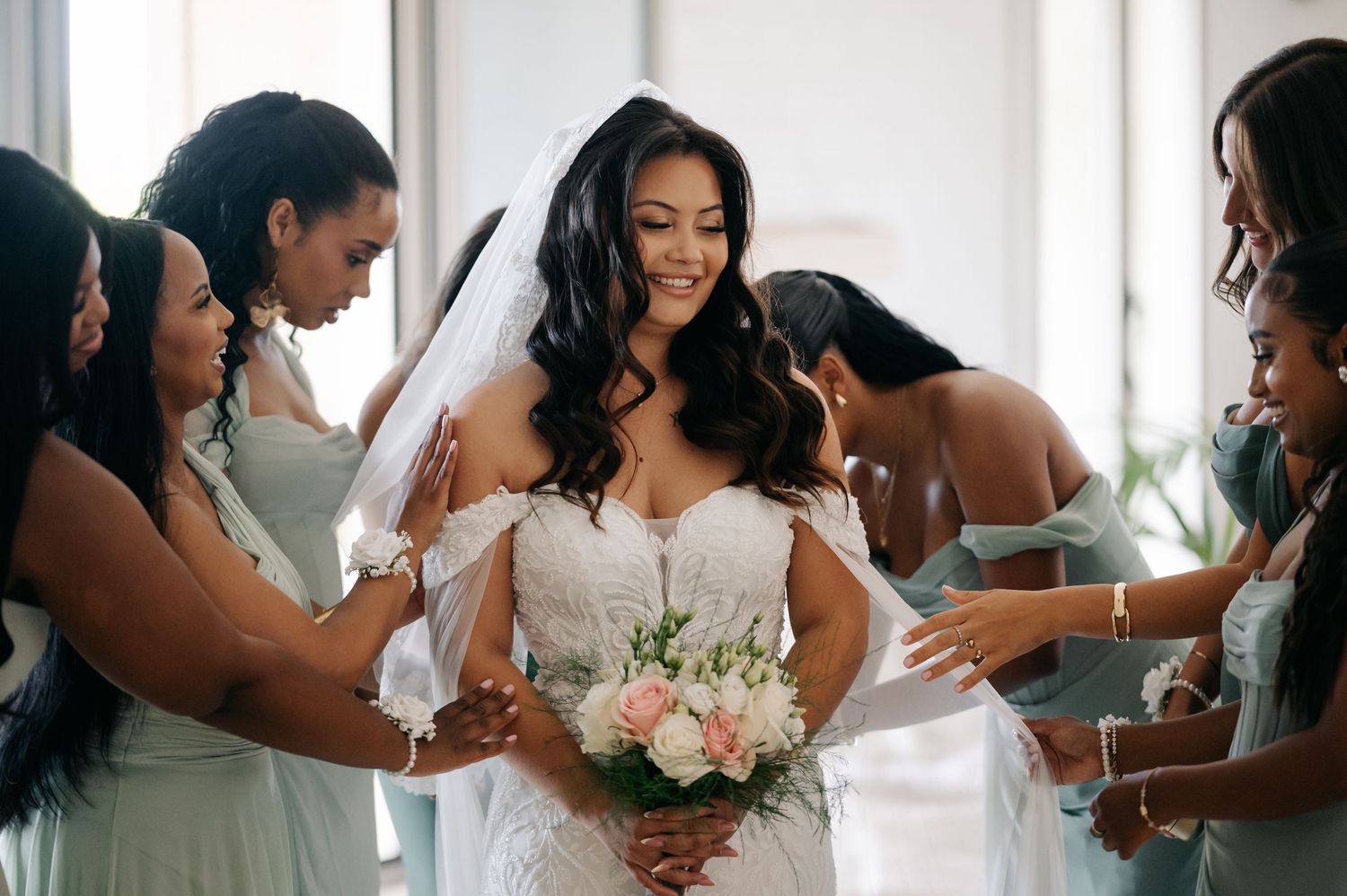 Bridesmaids in light blue dresses help adjust a bride's white wedding gown as she holds a pink and white bouquet.