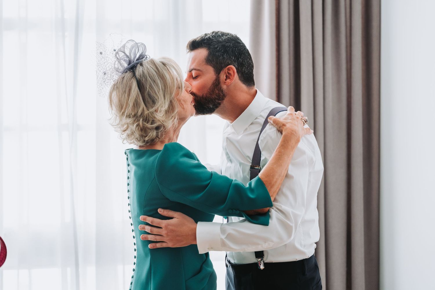 A couple in elegant attire shares a tender moment by a window with sheer curtains.